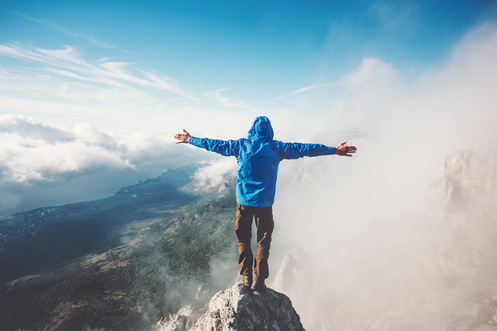 A man in a blue jacket stands at the peak of a mountain with his arms outstretched.